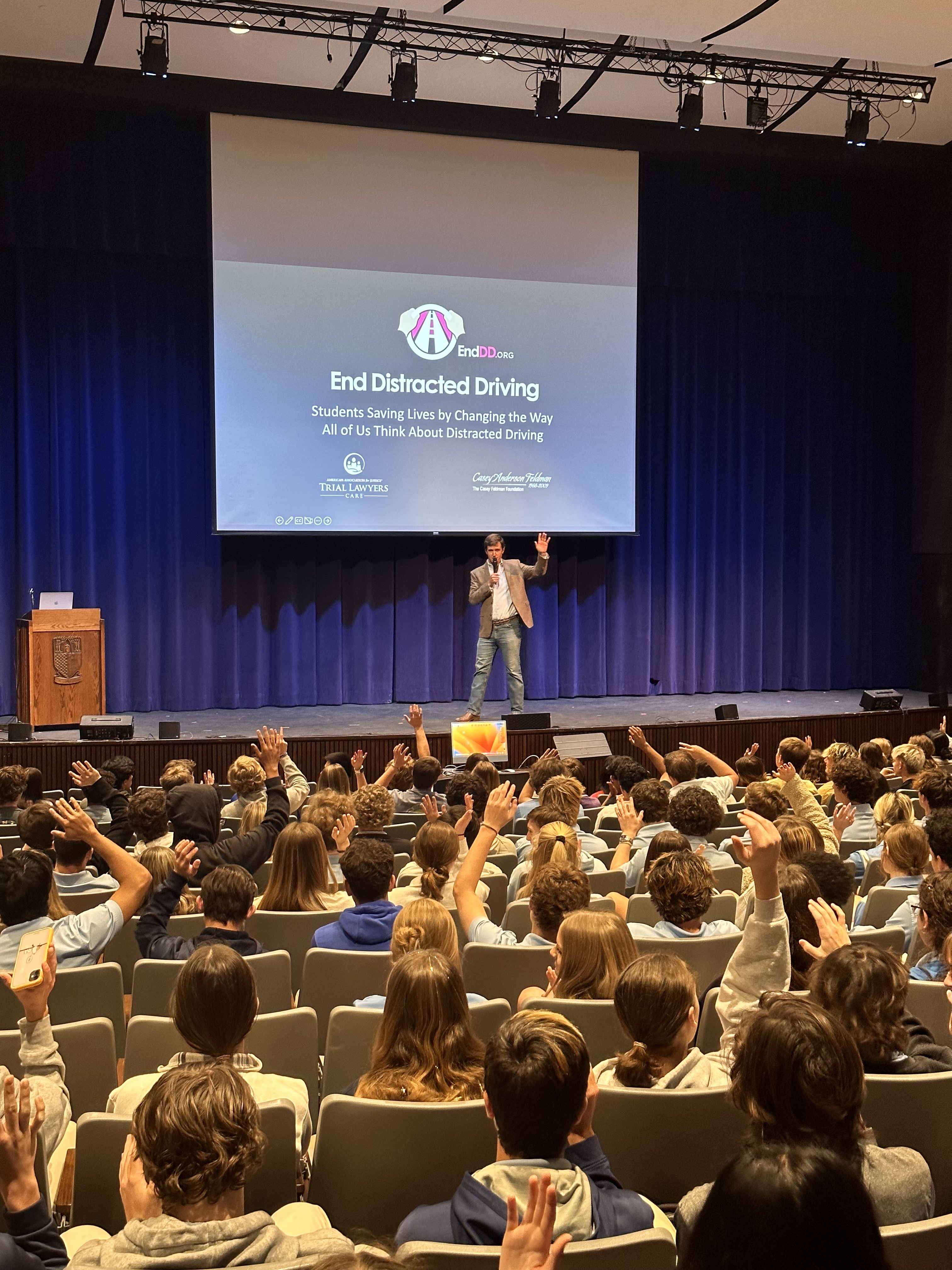 A photo of a man standing on an auditorium stage speaking to room of school age children who are seated and looking at the stage