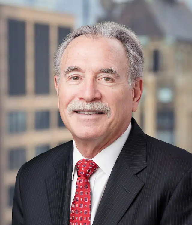 Dov Apfel against a blurry building backdrop wearing a black suit and red tie