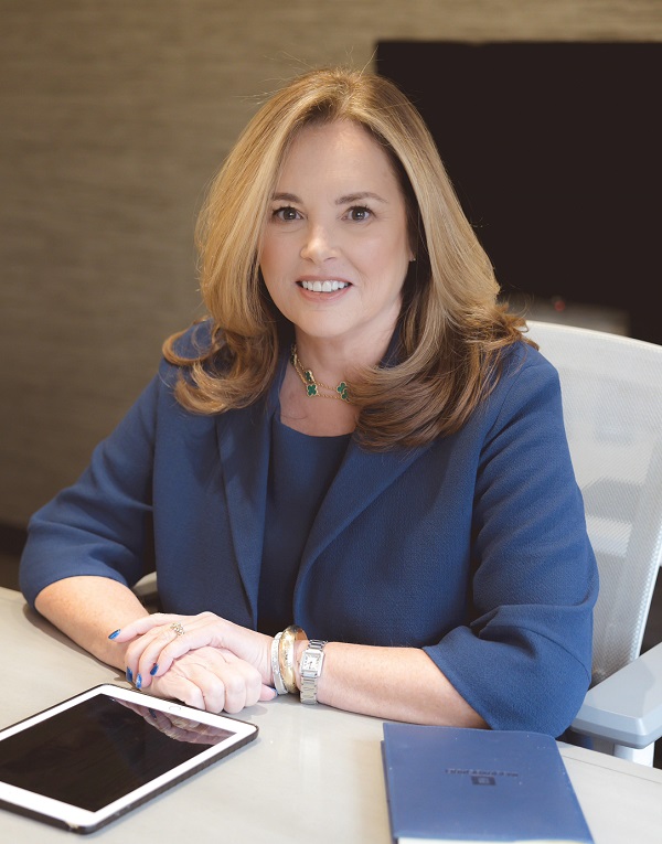 Marion Munley wearing a blue blazer and sitting in light colored chair and desk  with a book and ipad.