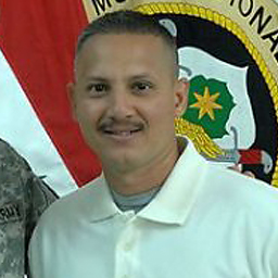 Picture of a man from the chest up with very short dark hair and mustache, wearing a white polo and standing in front of a marine corp flag.