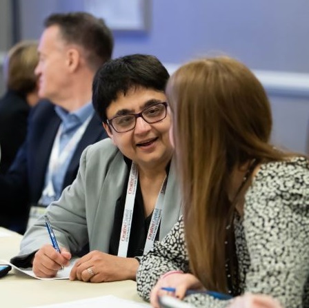 Two women speaking at a table with a male member sitting behind them.