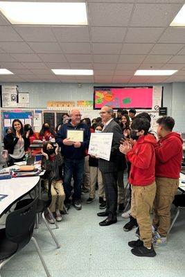 A photo of two men, one is holding an oversized check as they are surrounded by school age children 